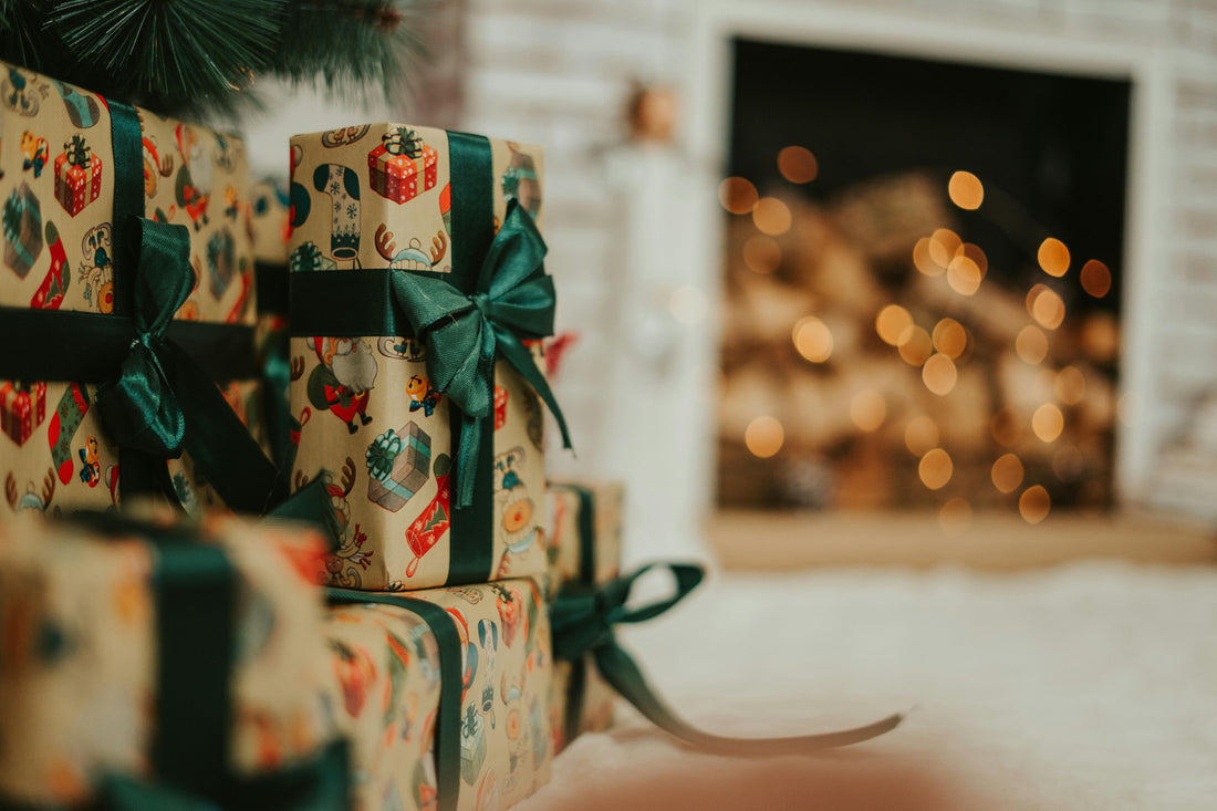 Christmas gifts wrapped in festive paper with green ribbon near a fireplace with blurred lights.