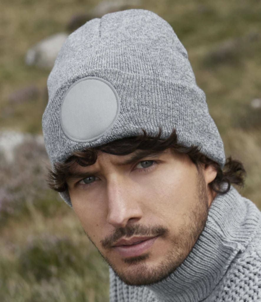 Man wearing a gray knit beanie with a circular pattern, standing in a natural setting.